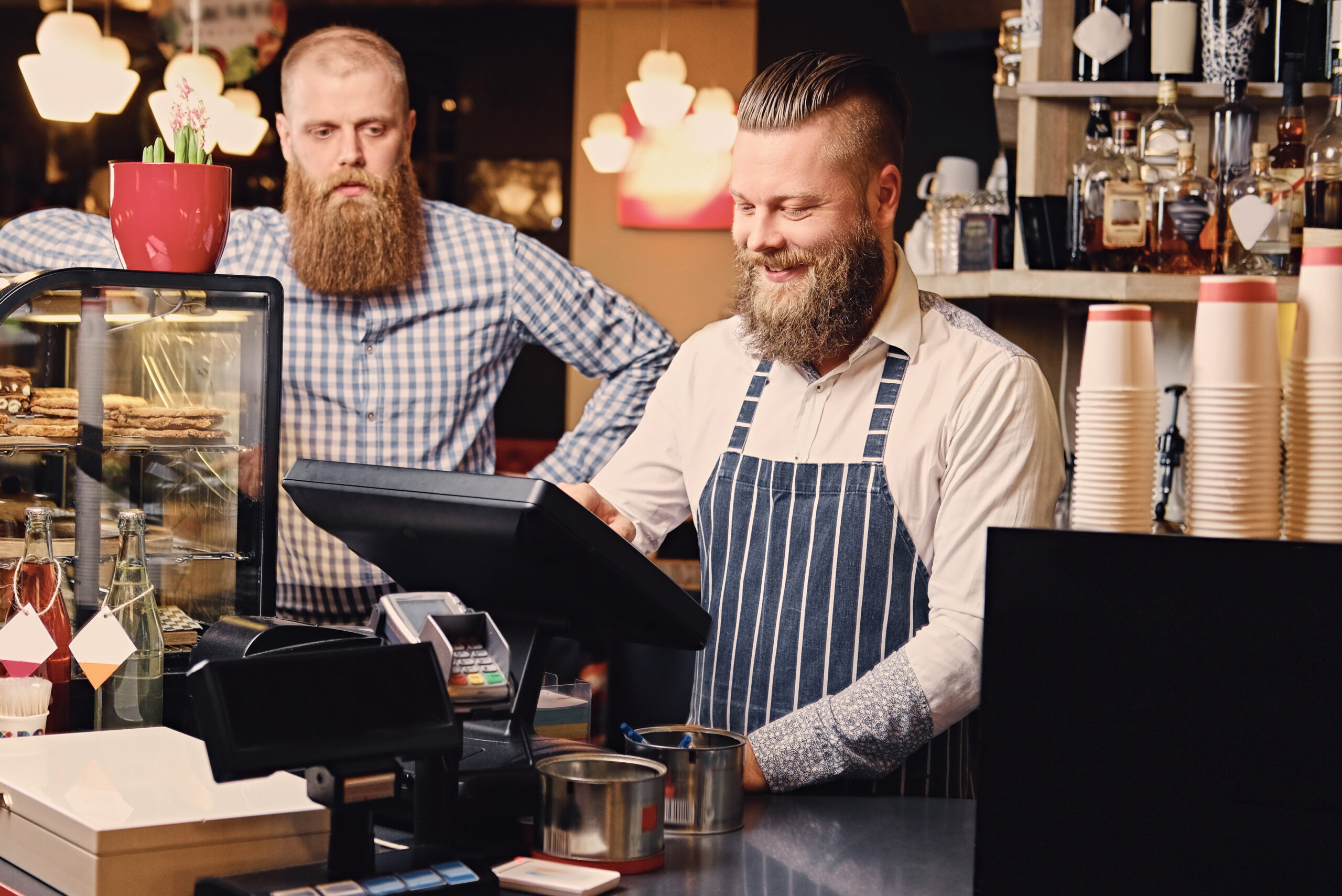 Positive bearded male at the counter using cash register in a coffee shop.